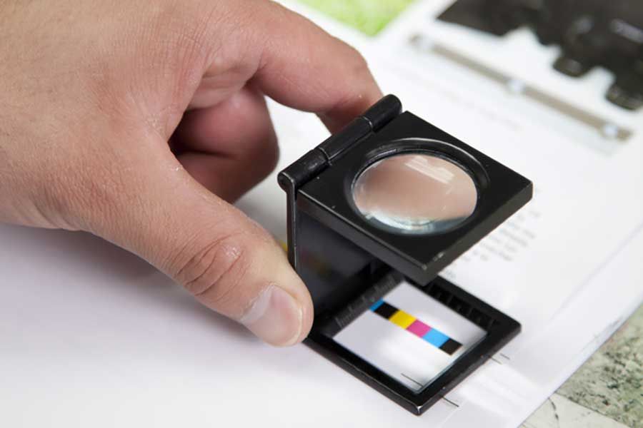 Hand holding magnifying loupe over printed colour chart, quality control inspection and surface analysis concept